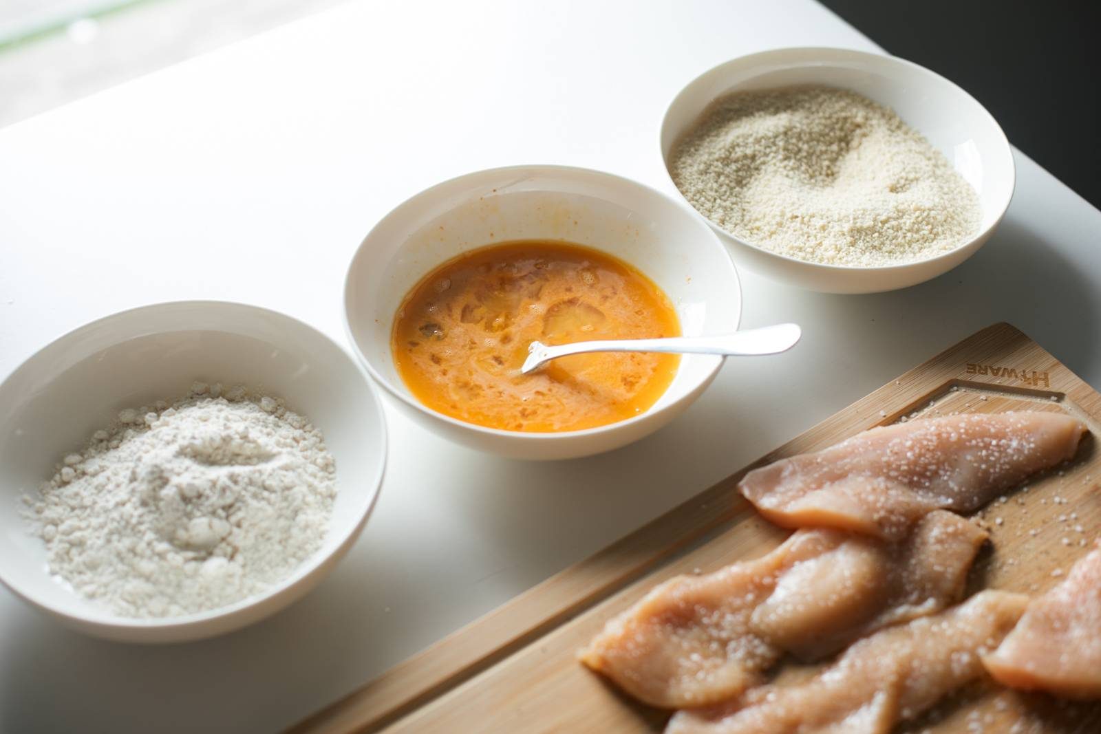 Flour, eggs, and breadcrumbs in bowls with chicken cutlets on a cutting board.